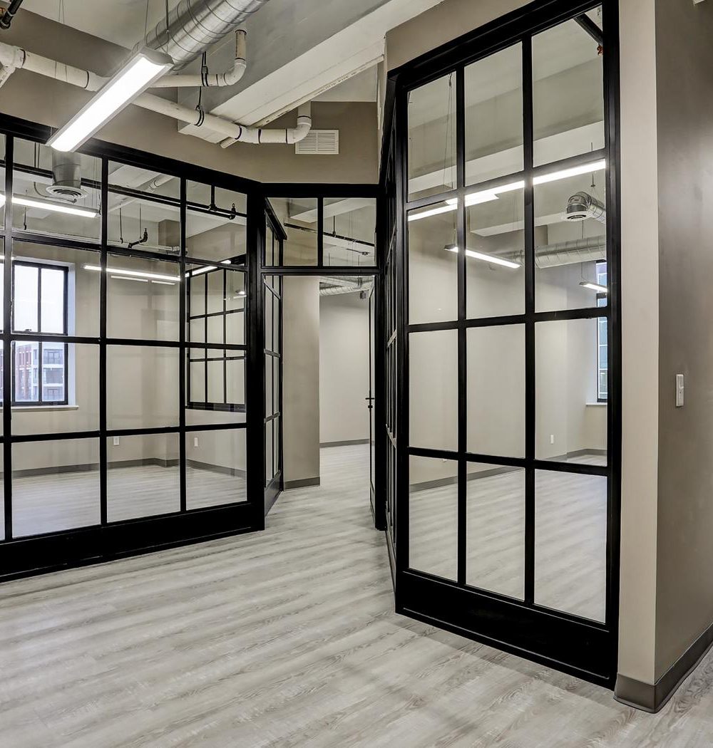 Vacant office with black-framed glass partitions, gray plank flooring, and exposed ceiling ducts.