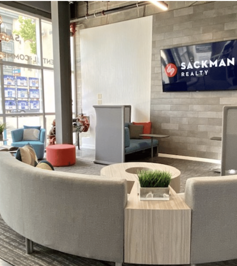 Furnished lobby lounge with curved gray seating, colorful accent chairs, and a Sackman Realty sign.