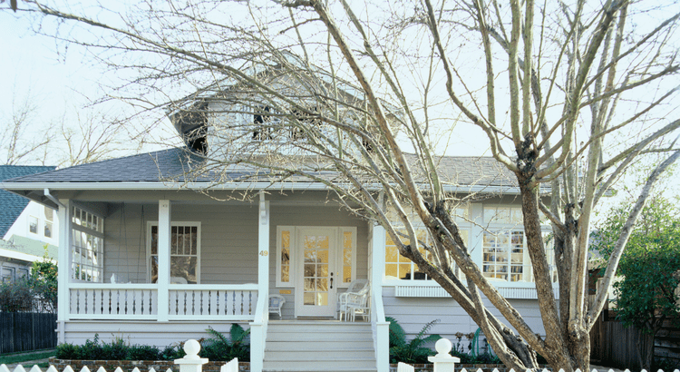 A quaint grey house with a gabled roof and front porch is surrounded by bare trees. White picket fence and porch chairs create a serene, cozy atmosphere.