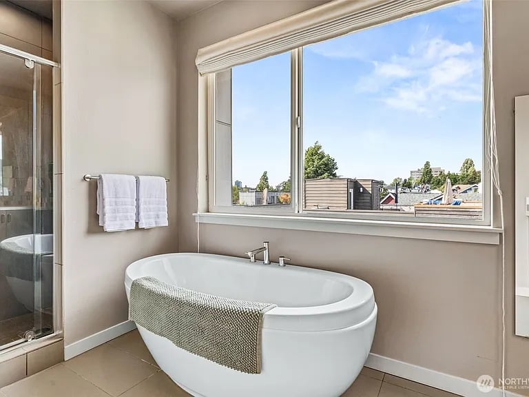 Bathroom with a freestanding soaking tub beneath a large window overlooking nearby rooftops and trees.
