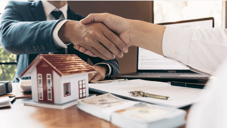 Real estate agents shake hands over sale documents beside a model house.