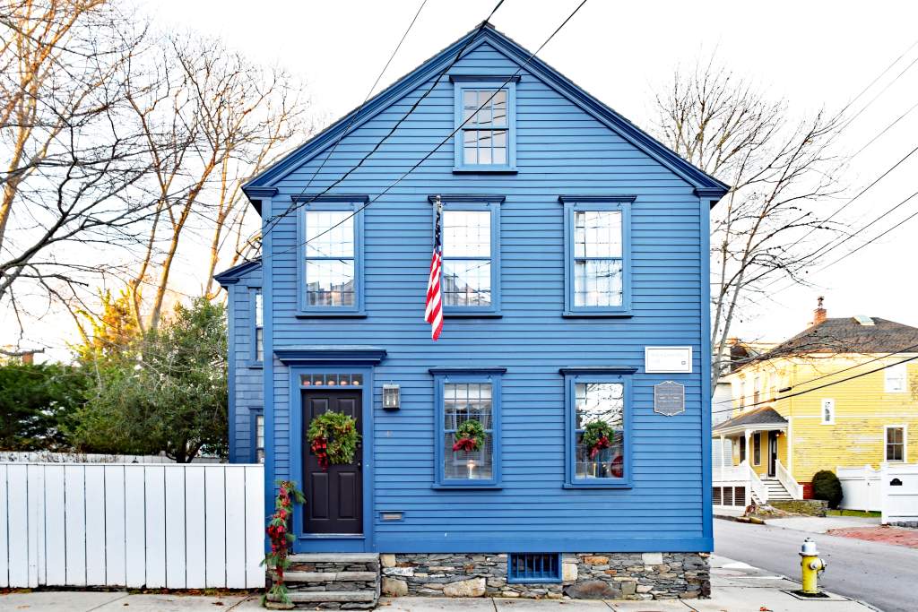 Blue two-story clapboard house with a stone foundation, American flag, and holiday wreaths.