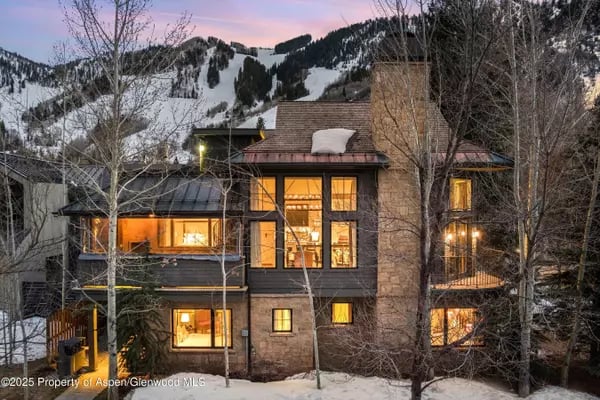 Mountain home with illuminated windows and a stone chimney, backed by snowy slopes at dusk.