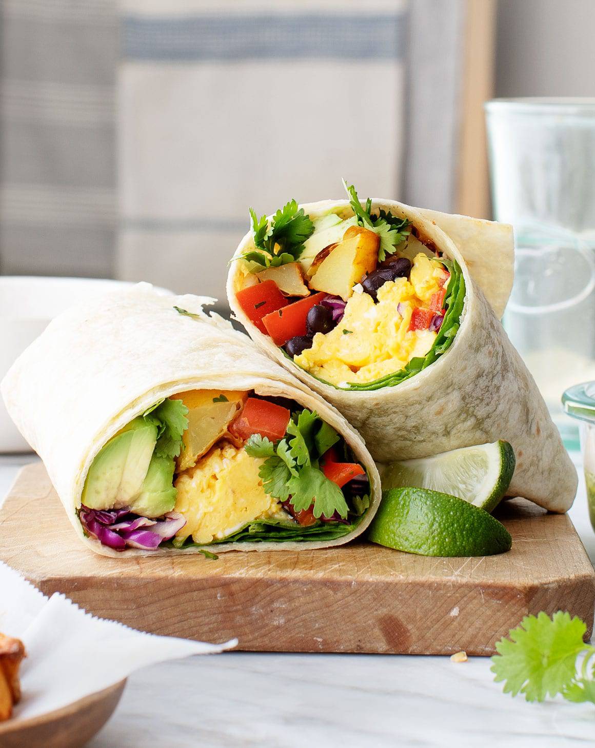Breakfast burrito halves filled with scrambled eggs, potatoes, beans, avocado, and greens, served with lime wedges.