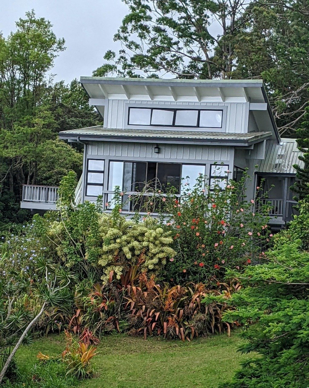 Modern gray house with broad angular roof, raised deck, and lush flowering garden.