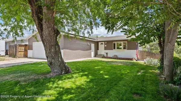 Single-story ranch home with an attached garage, shaded lawn, mature trees, and an American flag.