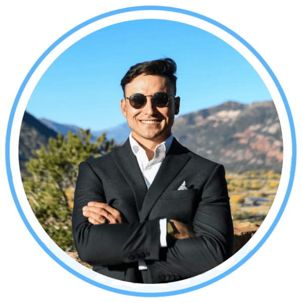 Man in a black suit and sunglasses poses with crossed arms against a mountain landscape.