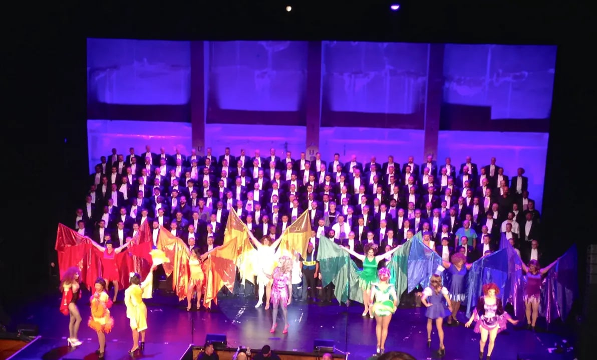 San Francisco Gay Men's Chorus on stage; over a hundred men in tuxedos stand behind performers in colorful drag and gowns holding large, sheer, brightly colored banners