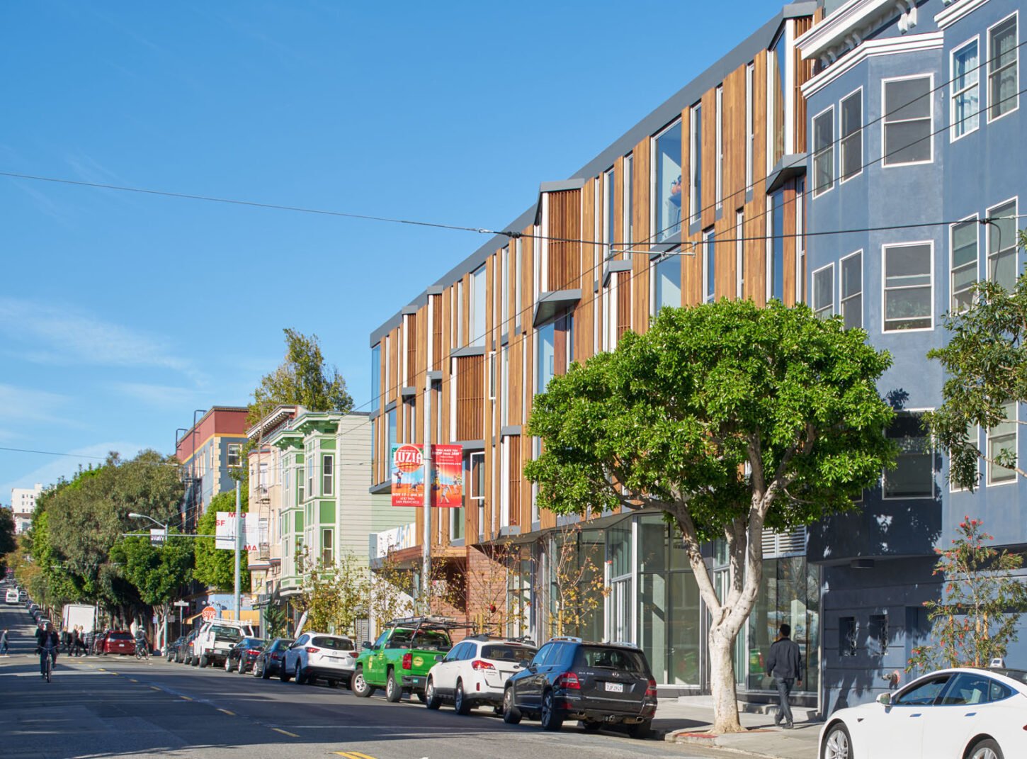 A street in Hayes Valley, San Francisco, featuring a mix of modern and traditional architecture. The modern building on the right has a wood-paneled facade with large windows, while adjacent structures showcase classic Victorian-style architecture. The street is lined with trees, parked cars, and small businesses, creating a vibrant urban atmosphere under a clear blue sky.