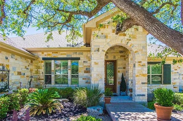 Stone house with an arched entry, garden beds, and a large shade tree in front.