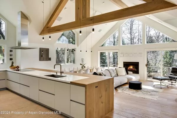Open-plan kitchen and living room with exposed timber beams, a fireplace, and large windows overlooking bare trees.
