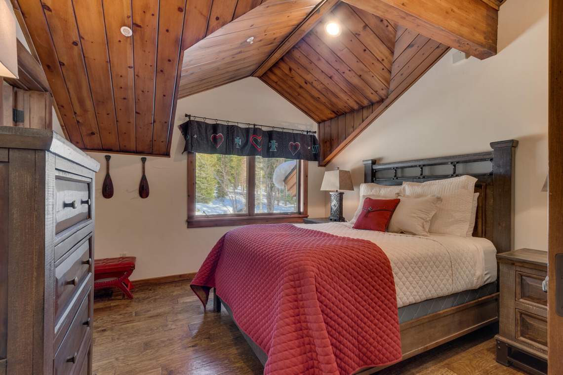Bedroom with a red quilt, vaulted wood ceiling, and window overlooking snow-dusted trees.