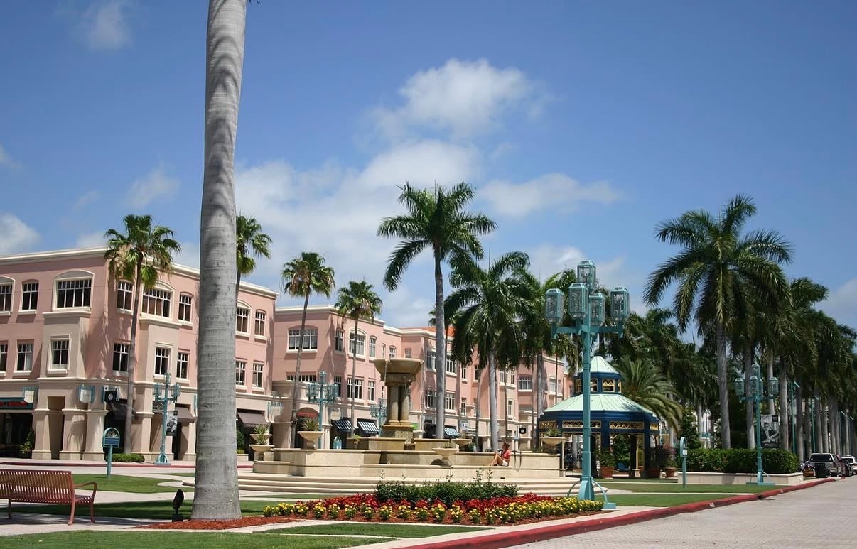Mizner Park plaza with palm trees, a fountain, colorful flower beds, and pink storefronts.