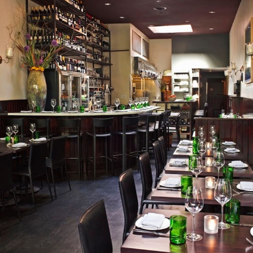 The interior of SPQR restaurant in San Francisco, featuring a slender dining space with a soaring ceiling, skylights, and an open kitchen. The atmosphere is kitschy and boisterous, with modern decor and tables set for guests.