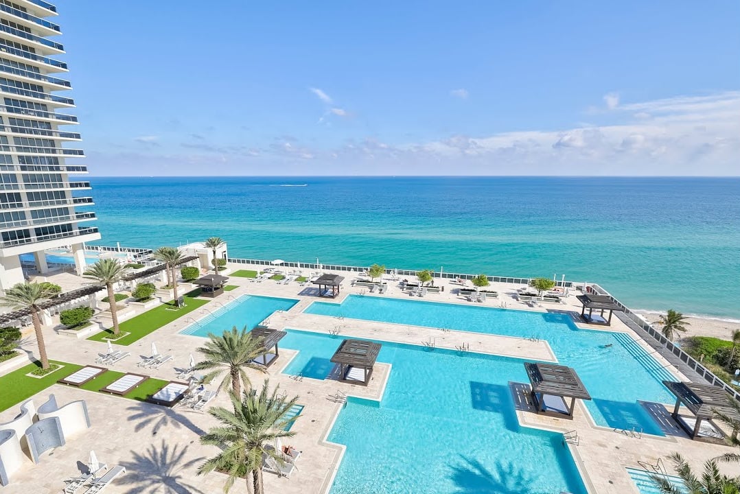 Oceanfront condominium pool deck with turquoise pools, palm trees, and the Atlantic Ocean beyond.