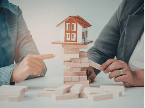 Two people handle blocks beneath a model house balanced on a wooden tower.