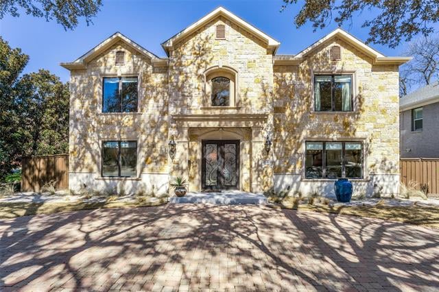 Two-story stone house with three gables, ornate double front door, and a tree-shadowed paved driveway.