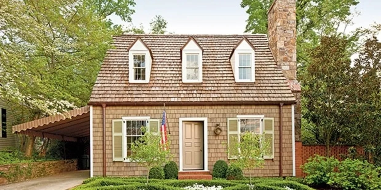 Shingle cottage with three dormers, a stone chimney, and a covered carport.