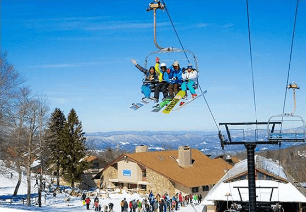 Skiers ride a chairlift above a resort lodge, with snowy mountains in the distance.