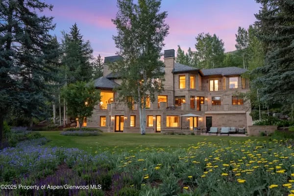Large stone-and-wood home with glowing windows, framed by evergreens and a flower-dotted lawn at dusk.