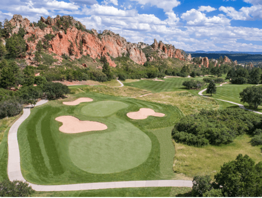 Golf course greens and sand bunkers below dramatic red rock formations.