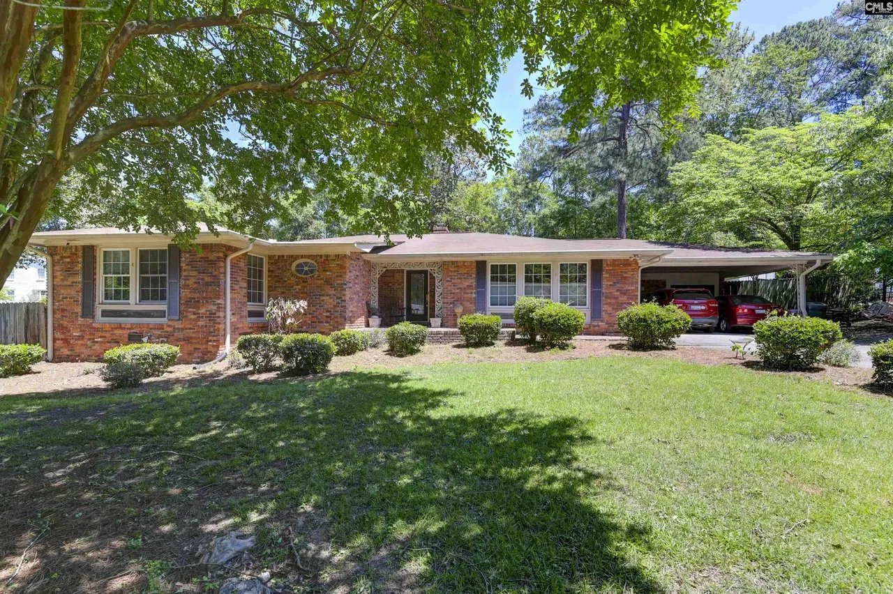 Red-brick ranch home beneath mature shade trees, with a broad lawn and covered carport.