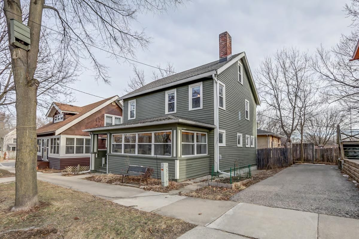 Green two-story house with an enclosed porch, brick chimney, and side driveway.