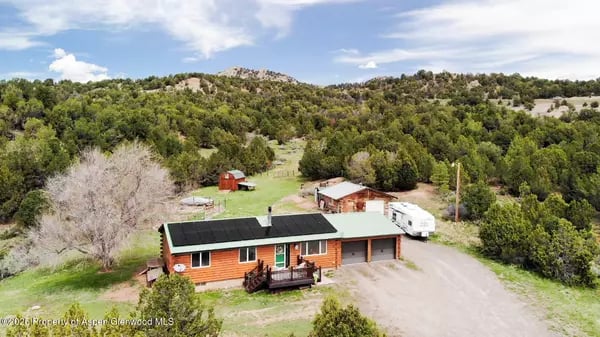 Aerial view of an orange-sided home with rooftop solar panels, an attached garage, and wooded hills beyond.