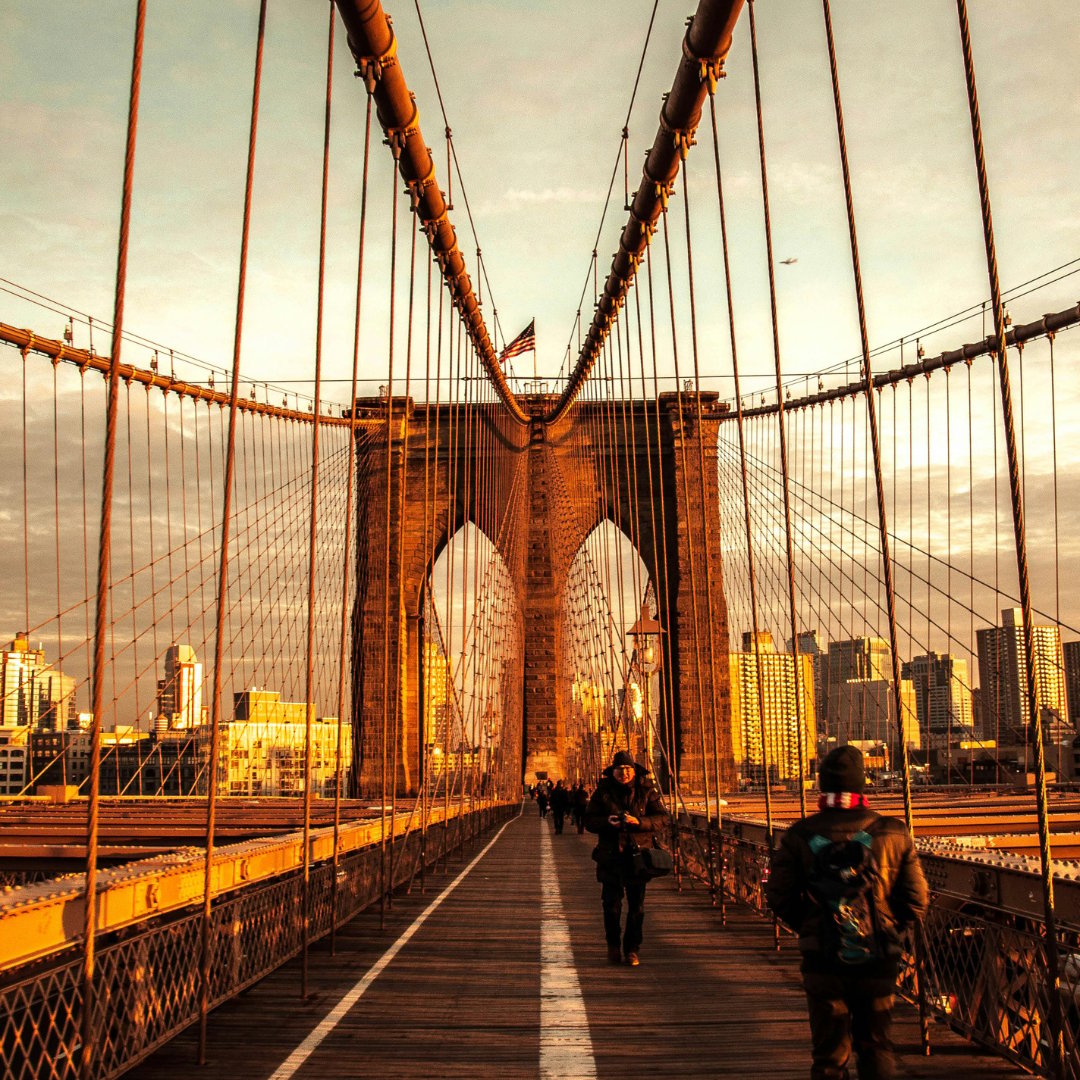 People walk across the Brooklyn Bridge toward its stone arch and the New York City skyline.