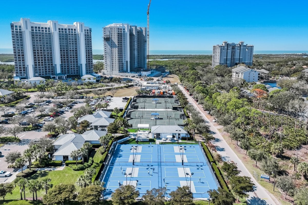 Aerial view over the Kalea Bay racket courts toward the towers, with one tower still under construction and a tower crane in place.