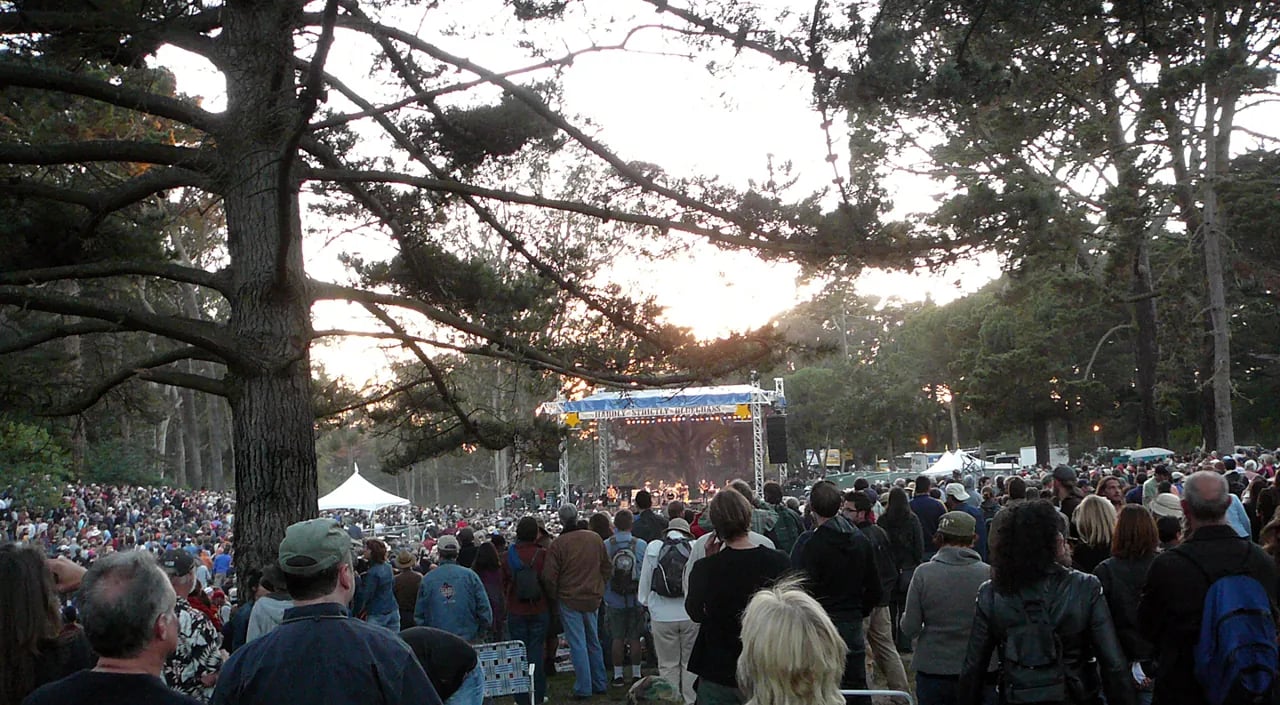 Crowd gathered at dusk in tree-lined meadow viewing distant stage with blue signage, with large tree silhouetted in foreground and sunset glow visible through branches