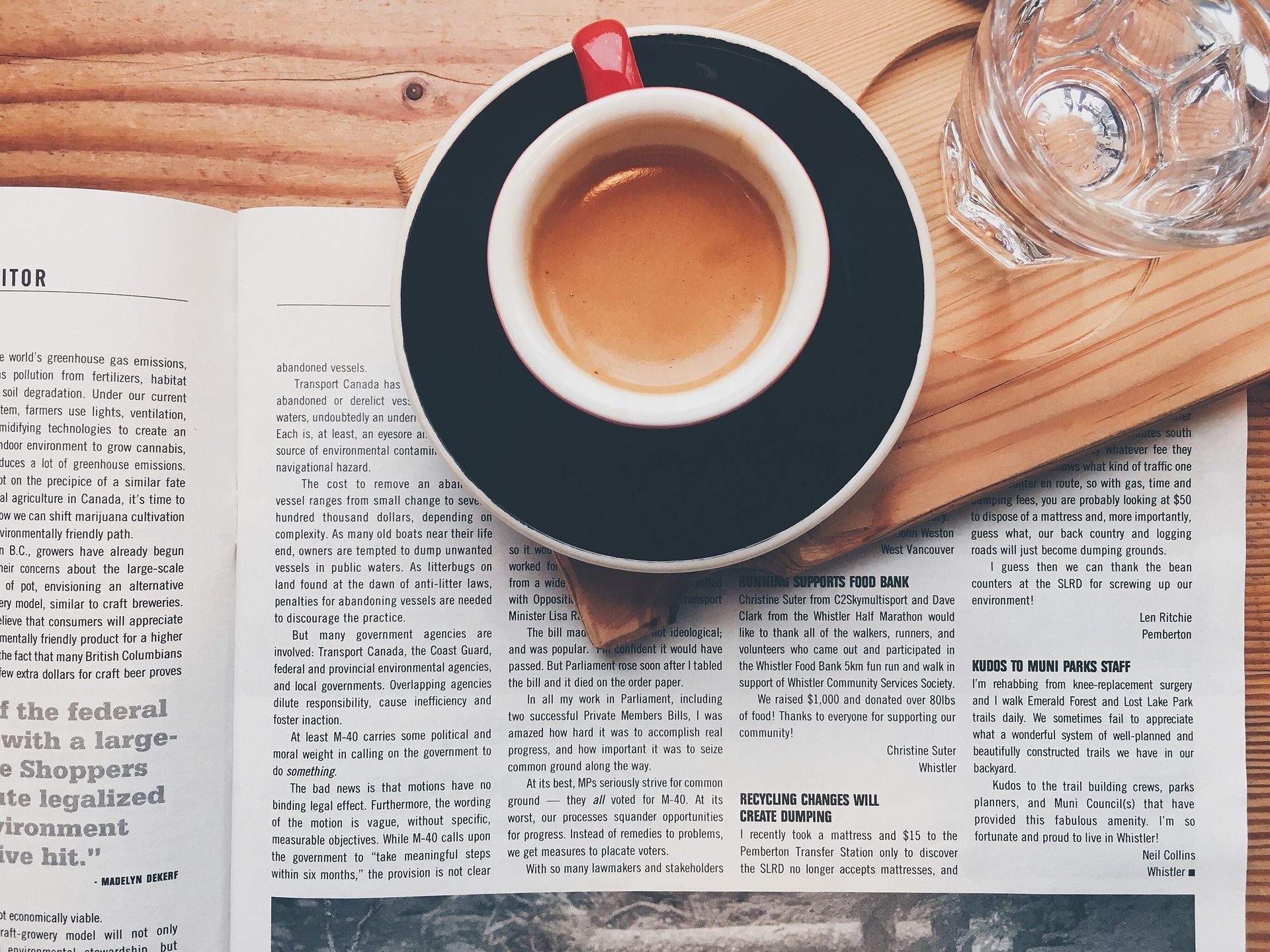 Cup of coffee on a dark saucer beside an open newspaper and clear glass.