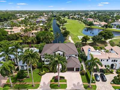 Aerial view of a large home beside a golf course and canal, with palm-lined landscaping.