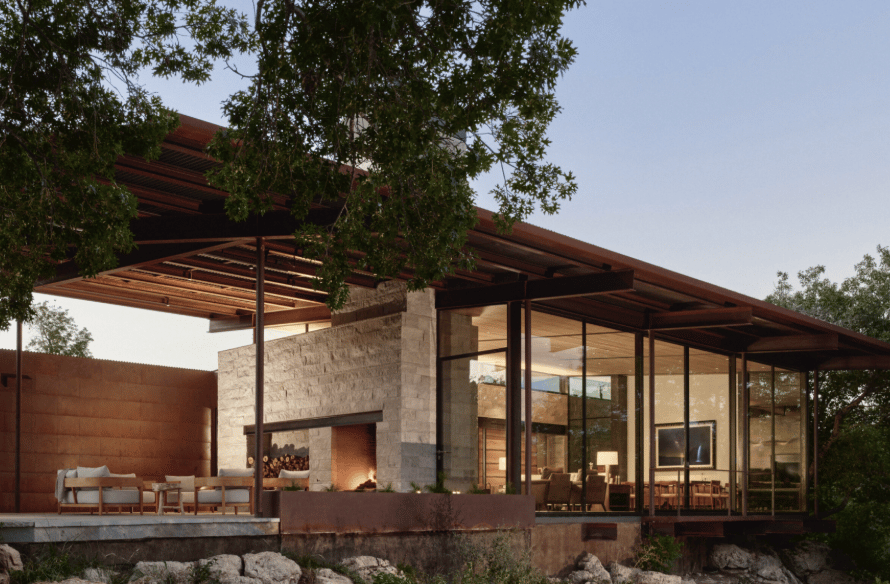 Modern home with glass walls, a covered patio, and an outdoor fireplace among trees.