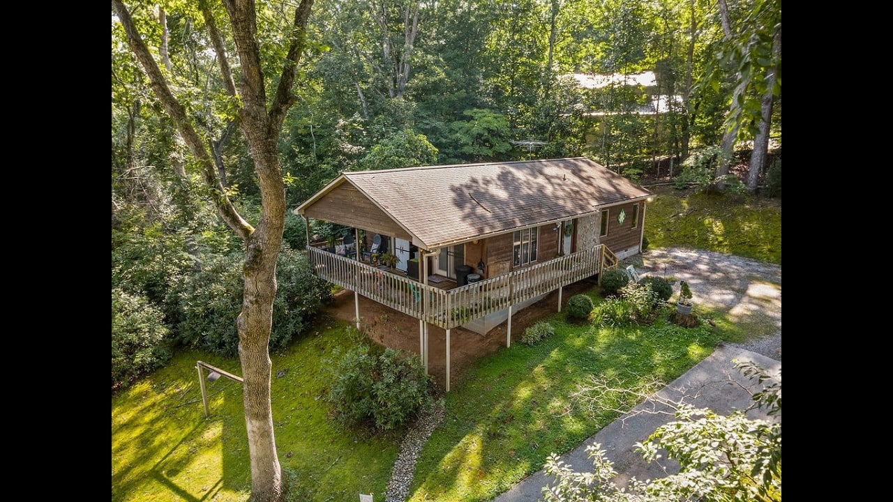 Aerial view of a brown home with a raised wraparound deck among mature trees.