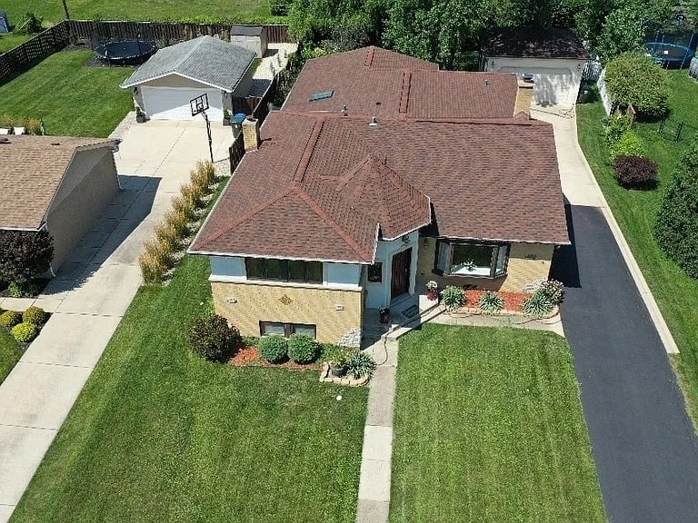 Aerial view of a single-story house with a reddish-brown roof, front lawn, and detached garage.