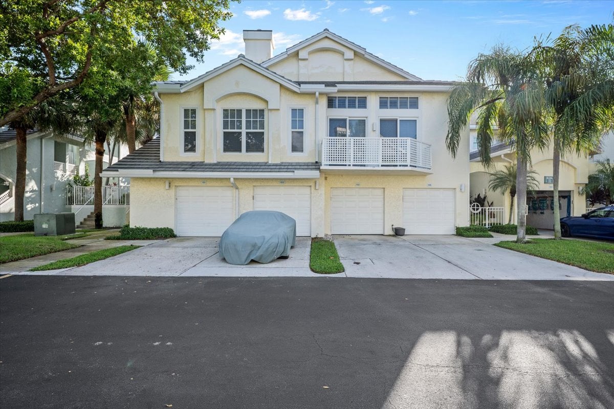 Cream-colored condominium exterior with a second-floor balcony, garage doors, and palm trees.