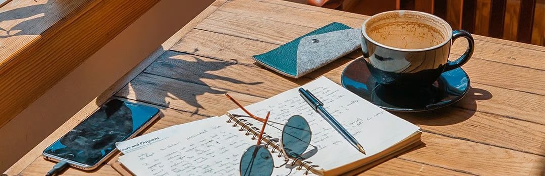 Open notebook, eyeglasses, smartphone, and coffee arranged on a sunlit wooden table.