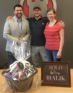 Three people celebrate a home closing beside a gift basket and framed SOLD sign.
