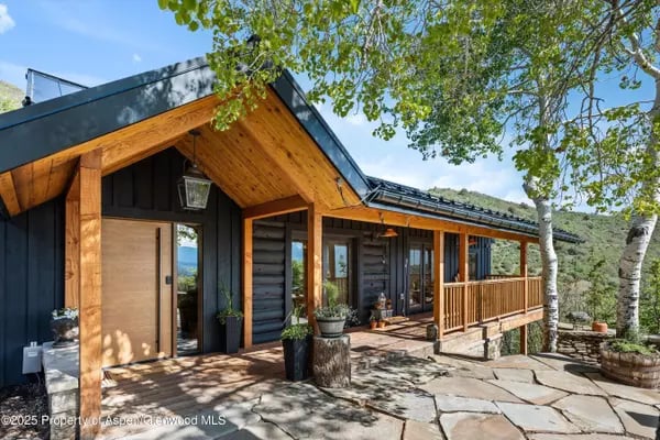 Modern mountain home with a timber-framed porch and stone patio beneath mature trees.