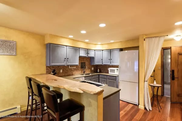 Kitchen with gray cabinets, a large stone breakfast bar, dark stools, and wood flooring.