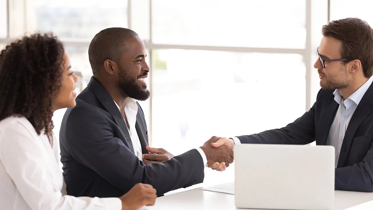A couple shakes hands with a professional across a desk in a bright office.