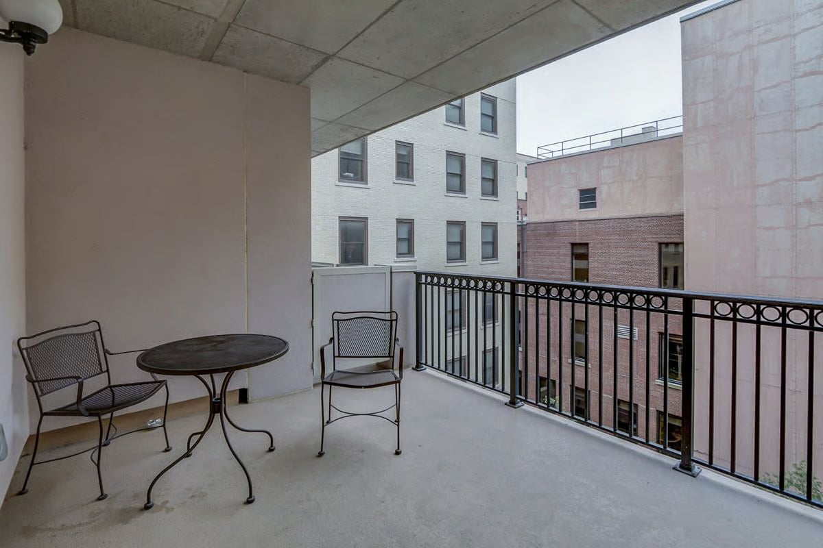 Covered apartment balcony with a metal railing, patio table and chairs, and neighboring buildings.