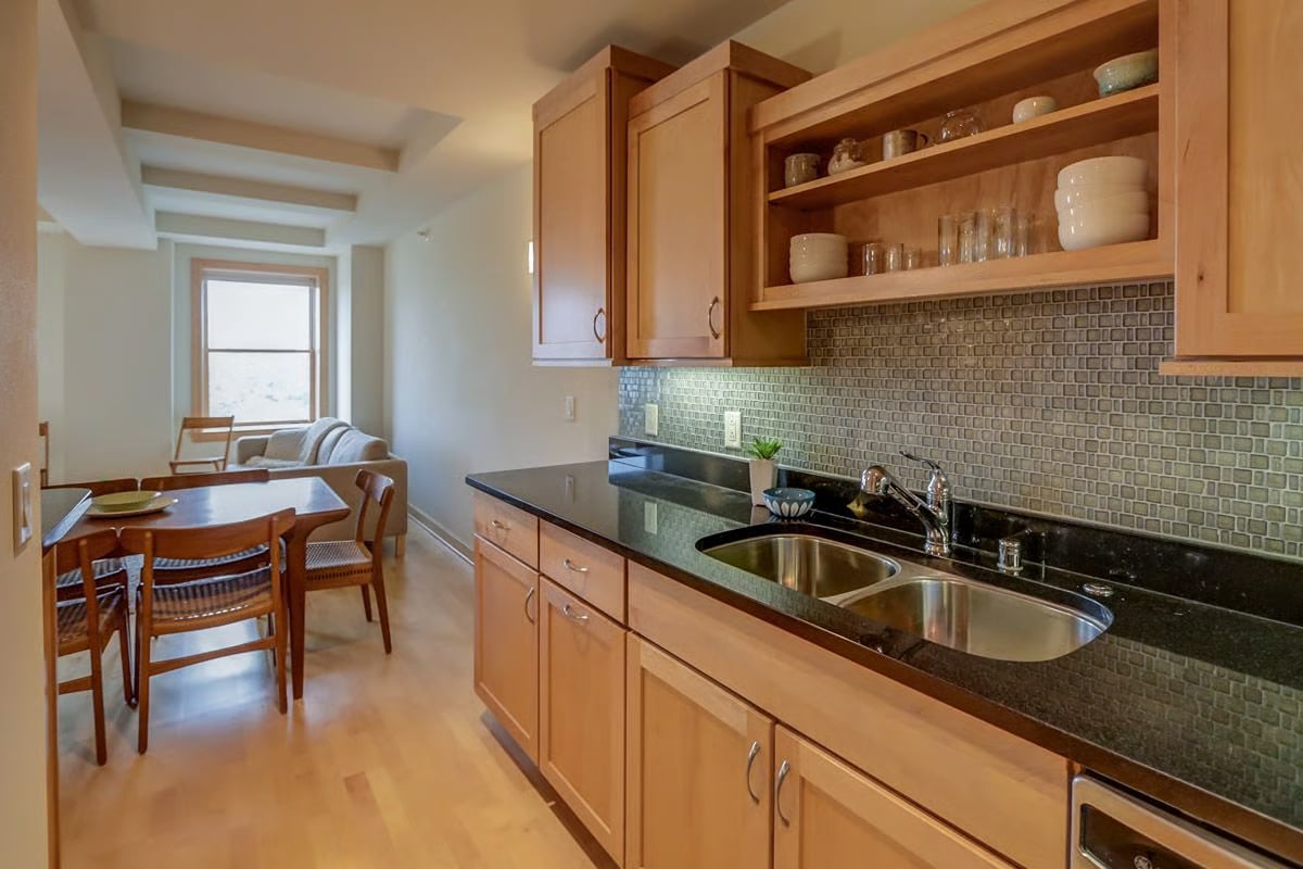 Kitchen with wood cabinets, open shelving, dark counters, and a double sink, opening onto a dining area.