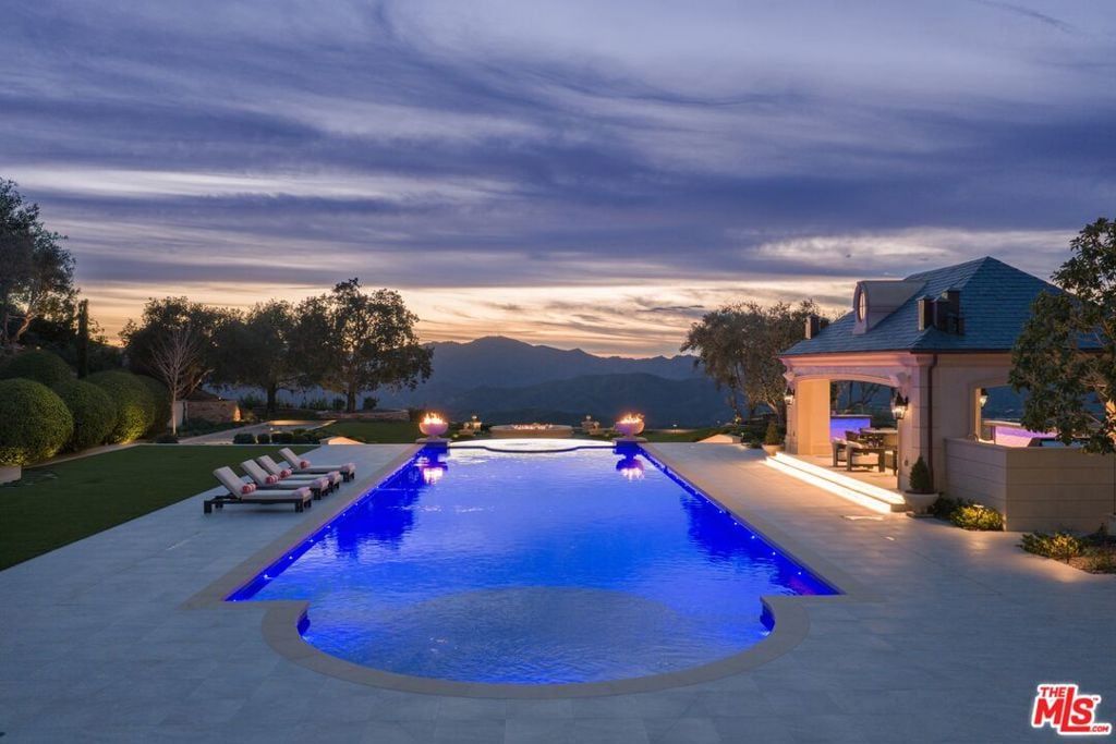 Backyard pool glowing at dusk, with lounge chairs, a covered seating area, and mountains beyond.