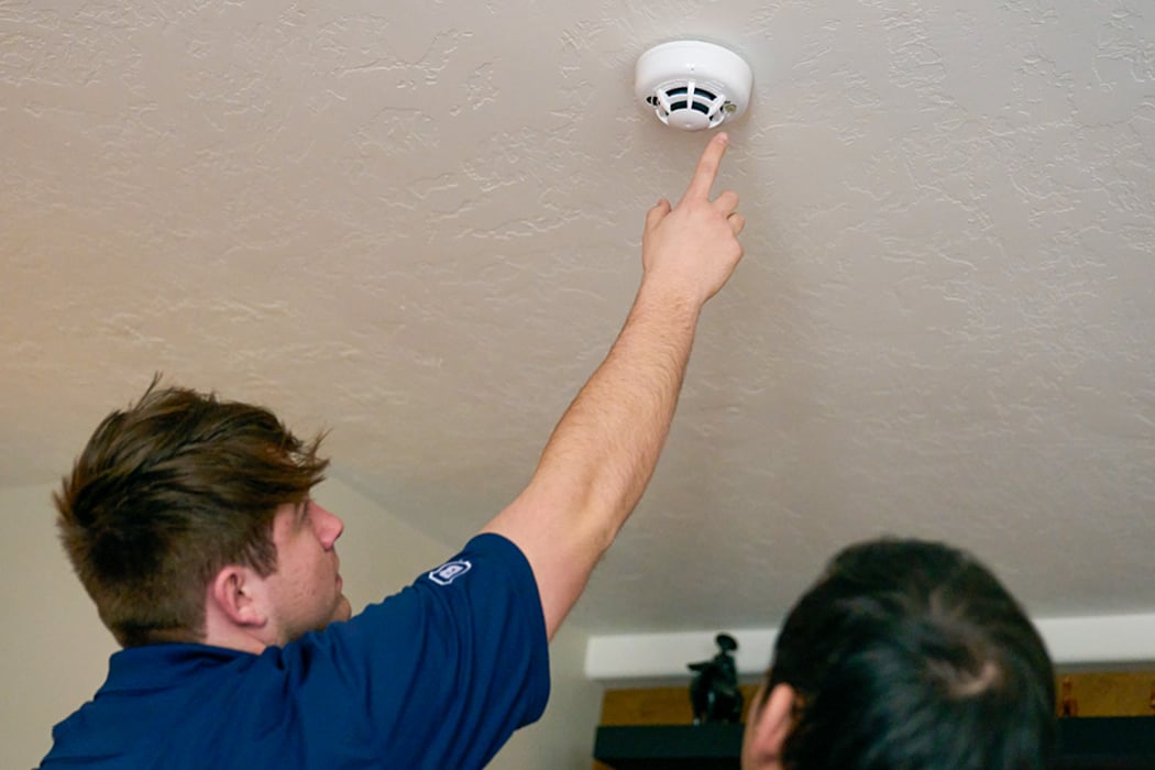 A man tests a ceiling-mounted smoke detector as another person watches.