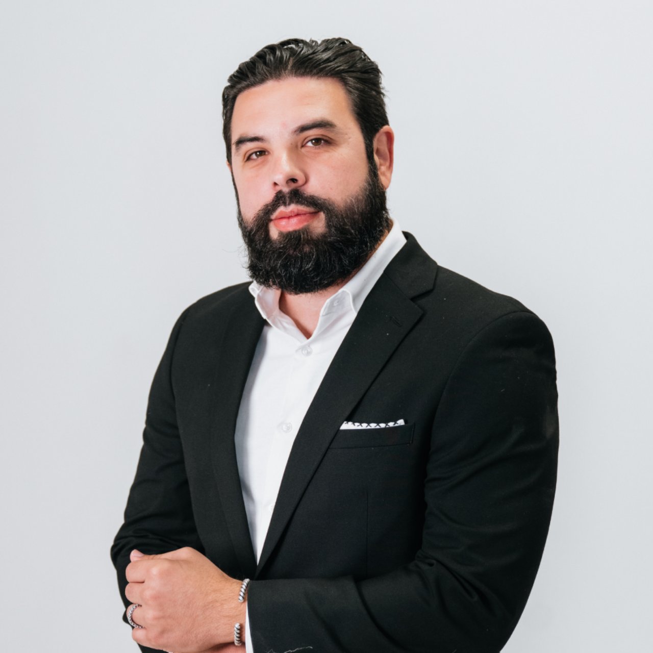 Bearded man in a black suit and white shirt, arms folded against a light gray background.