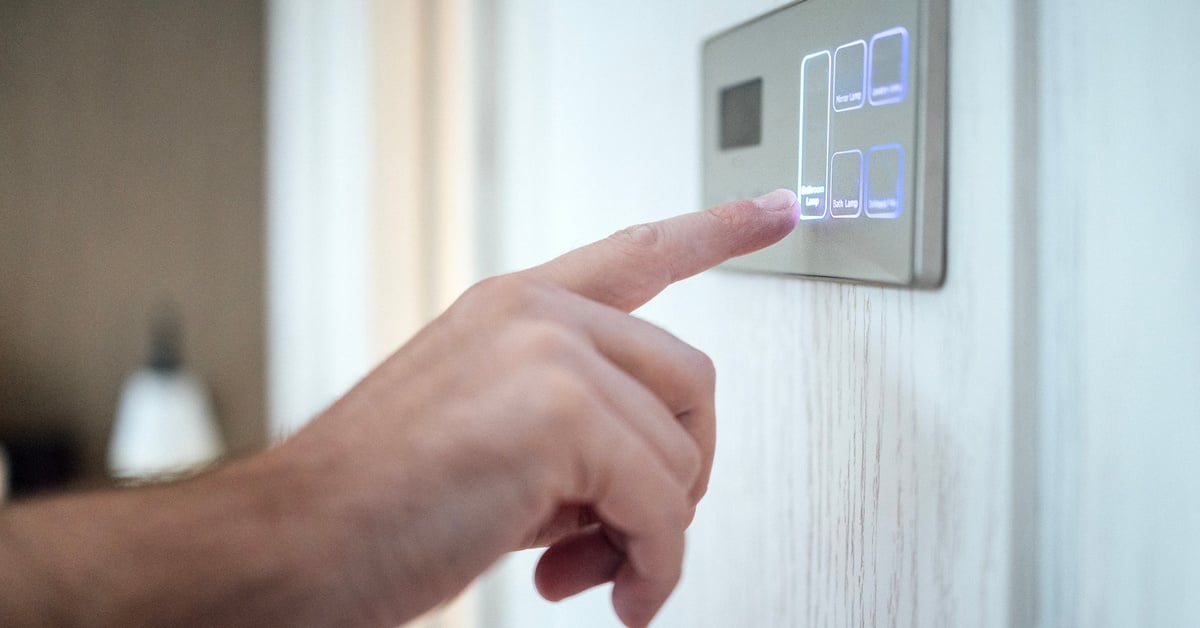 A close-up of a person touching a modern home control panel on a wall with an illuminated touch screen.