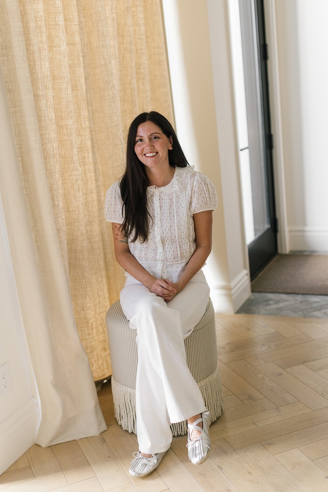 Danielle Tumanov seated on a fringed ottoman beside a window.