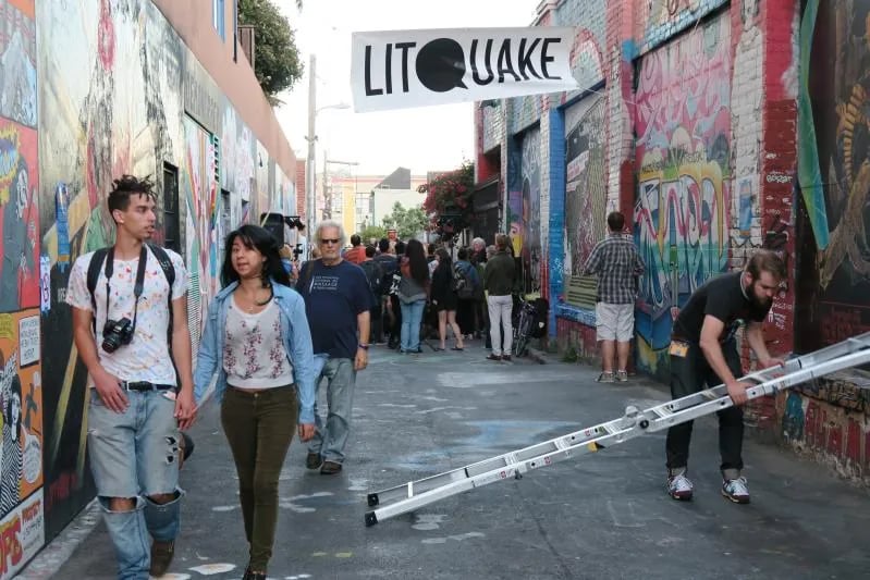 A crowd of people walking down a narrow urban alleyway covered in colorful murals and graffiti, beneath a hanging black and white banner that reads 